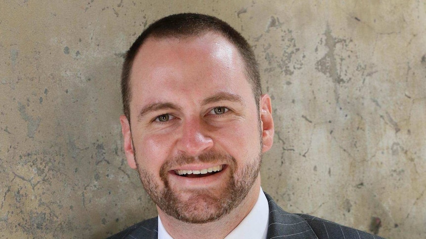 Andrew Bragg wears a suit and leans against a concrete wall