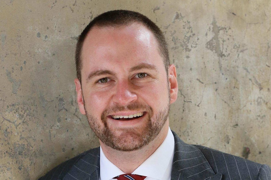 Andrew Bragg wears a suit and leans against a concrete wall