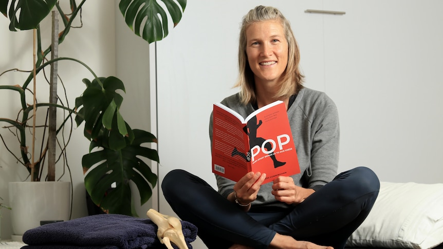 A smiling young woman with shoulder-length blonde hair holds a book with red cover, sits cross-legged, large plant in white pot.