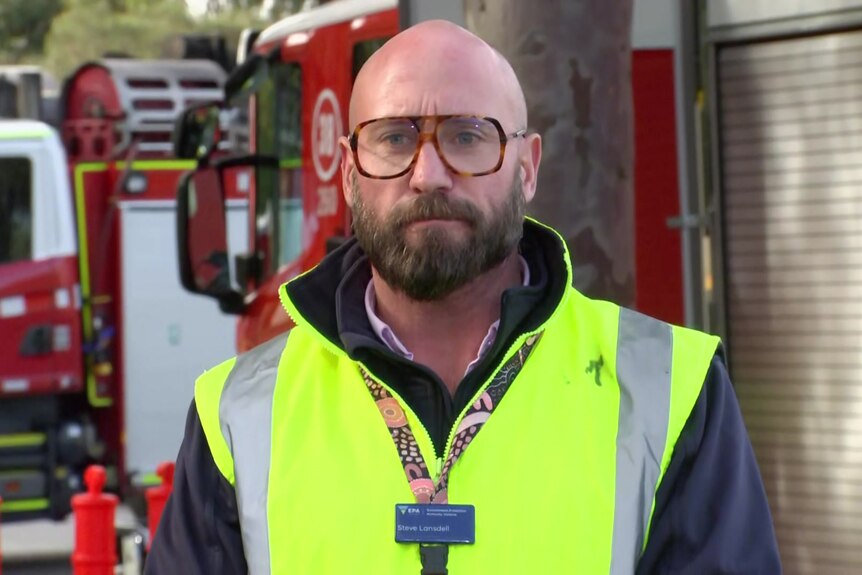Steve Lansdell wears a yellow fluro vest over a dark jacket and stands near two red fire trucks.