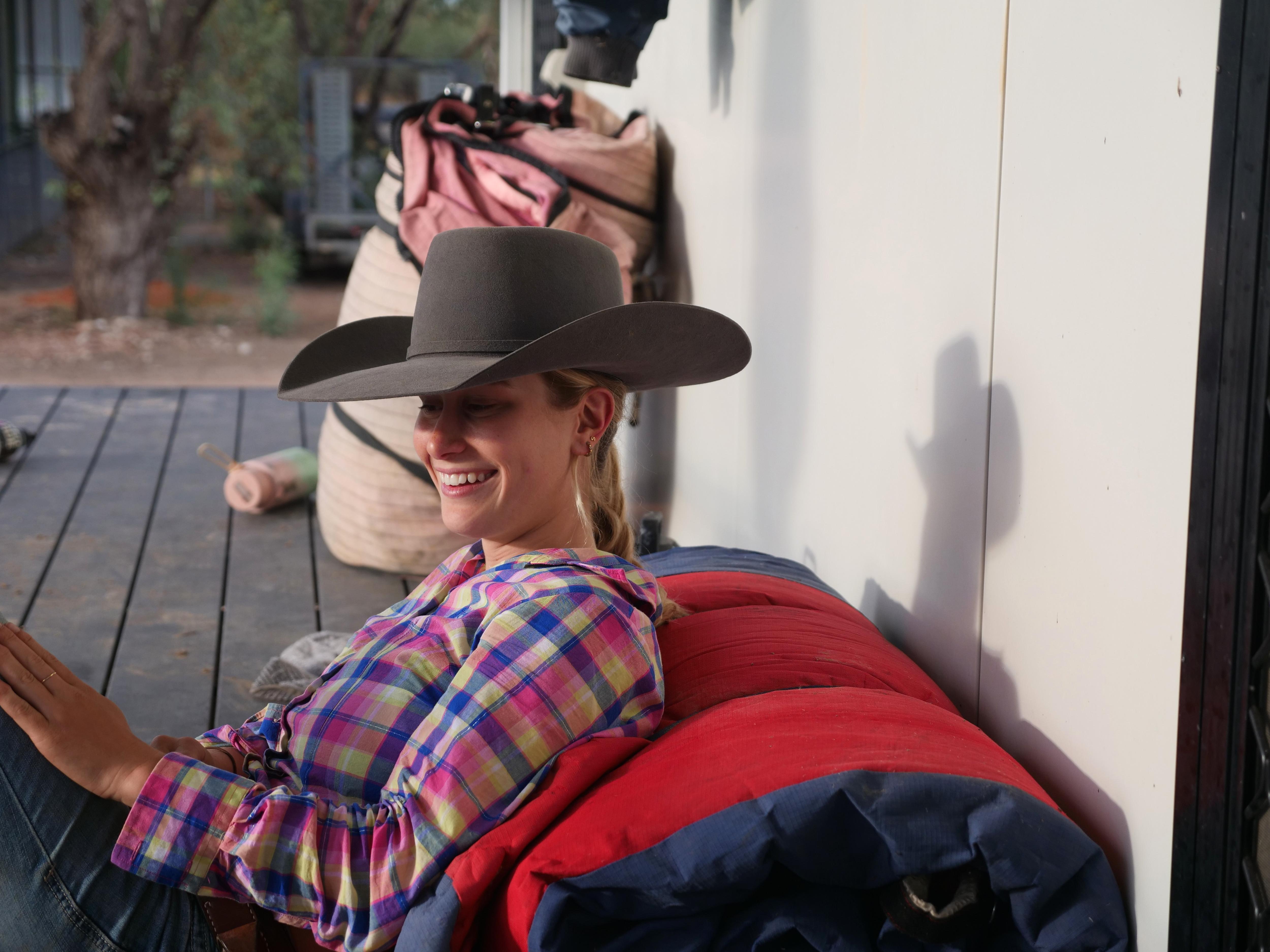 Sophie Bruce sentada em um terraço apoiada em uma bota sorrindo com um grande chapéu. 