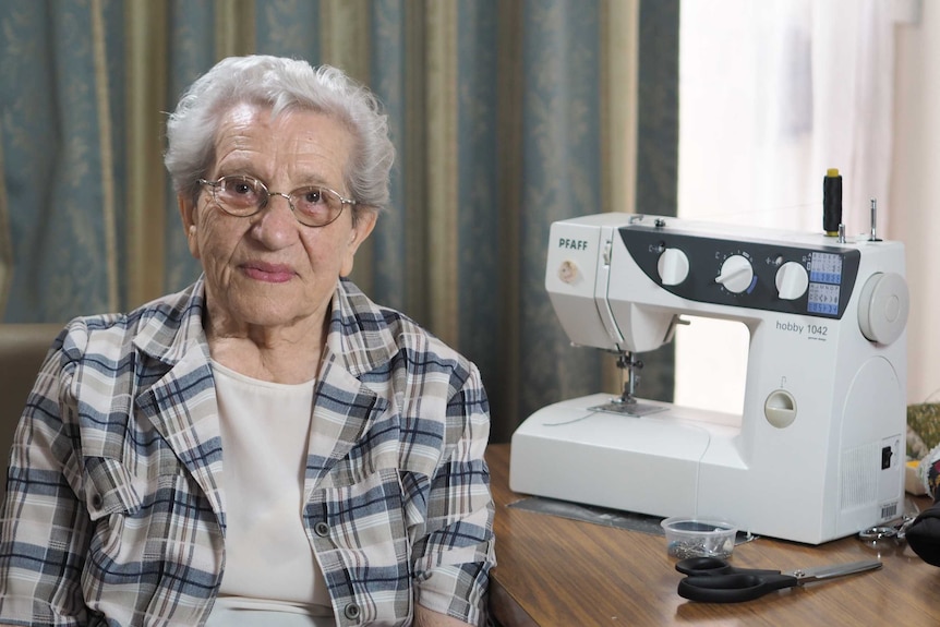 An elderly woman wearing glasses sitting next to a sewing machine
