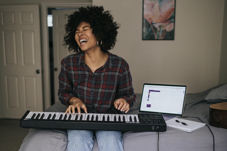 A woman sings while playing the keyboard, sitting on a bed. 