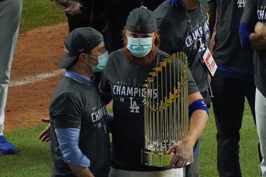 A Los Angeles Dodgers player, wearing a face mask, holds the World Series trophy on the playing field.
