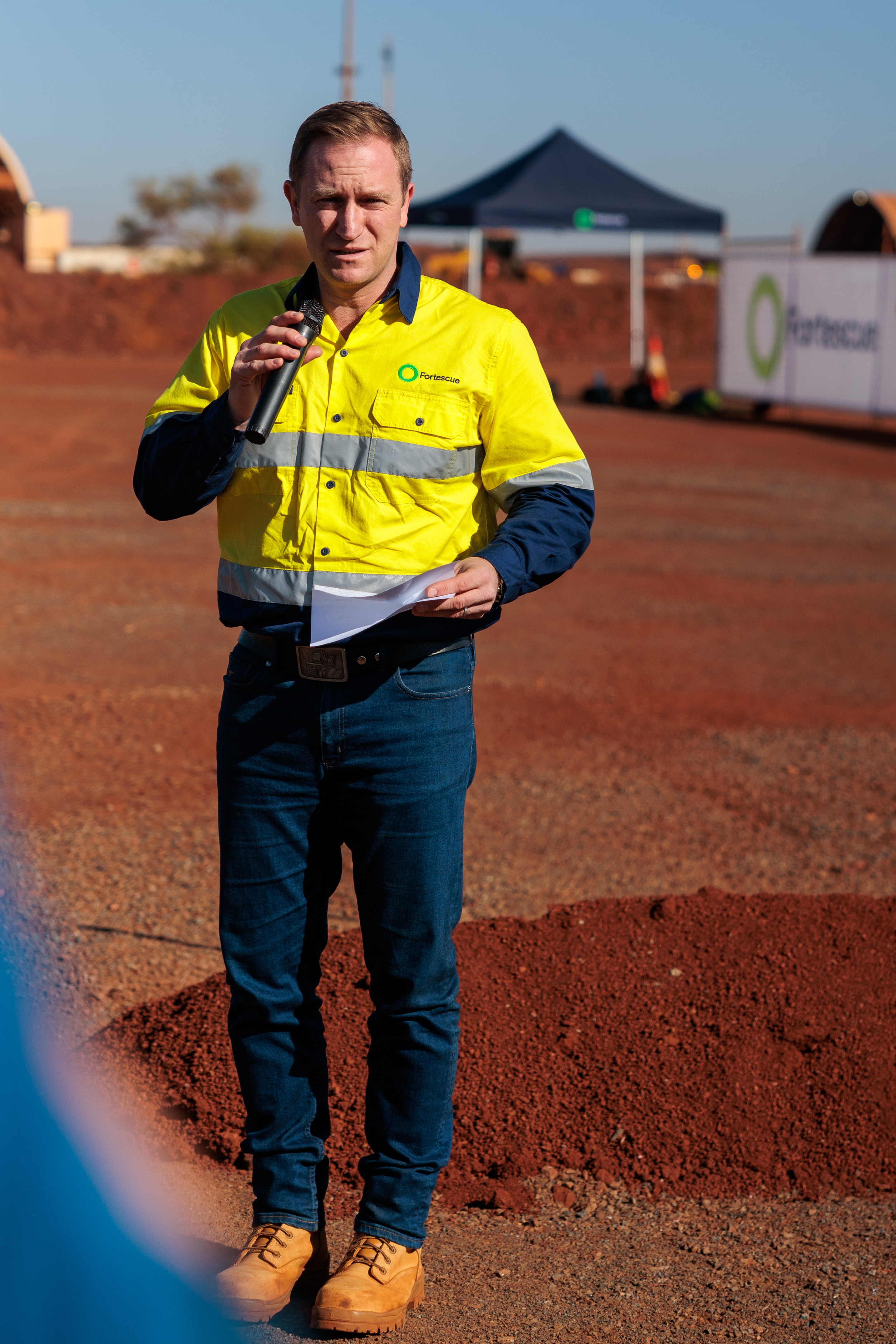 Um homem vestido de alta visibilidade segurando um microfone, dirigindo-se a uma multidão em um evento da Fortescue