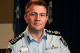 AFP Commissioner Reece Kershaw, in a uniform with flags hanging behind him in a room, looks seriously at the camera.