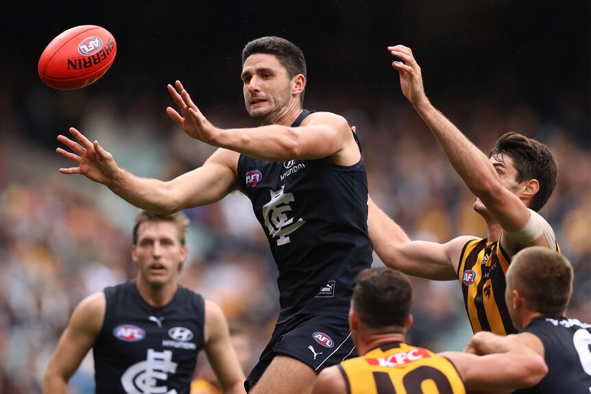 A tall AFL player in a Carlton jumper reaches out with his hands to grab the ball as a Hawthorn player grabs him from behind.