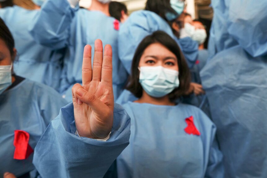 A woman wearing blue scrubs and a mask and a red ribbon holds up three fingers with a crowd of people in the background.