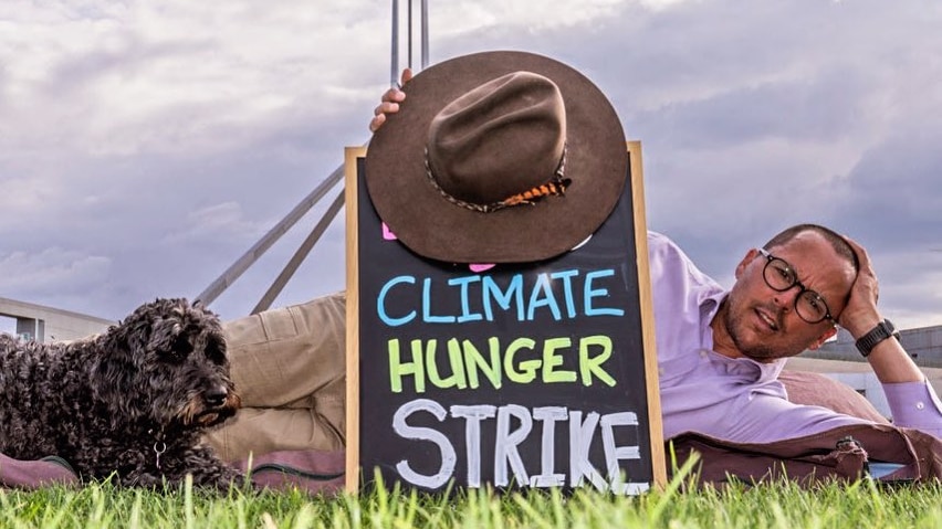a man with a sign saying "climate hunger strike" is laying on the lawn in front of Parliament House