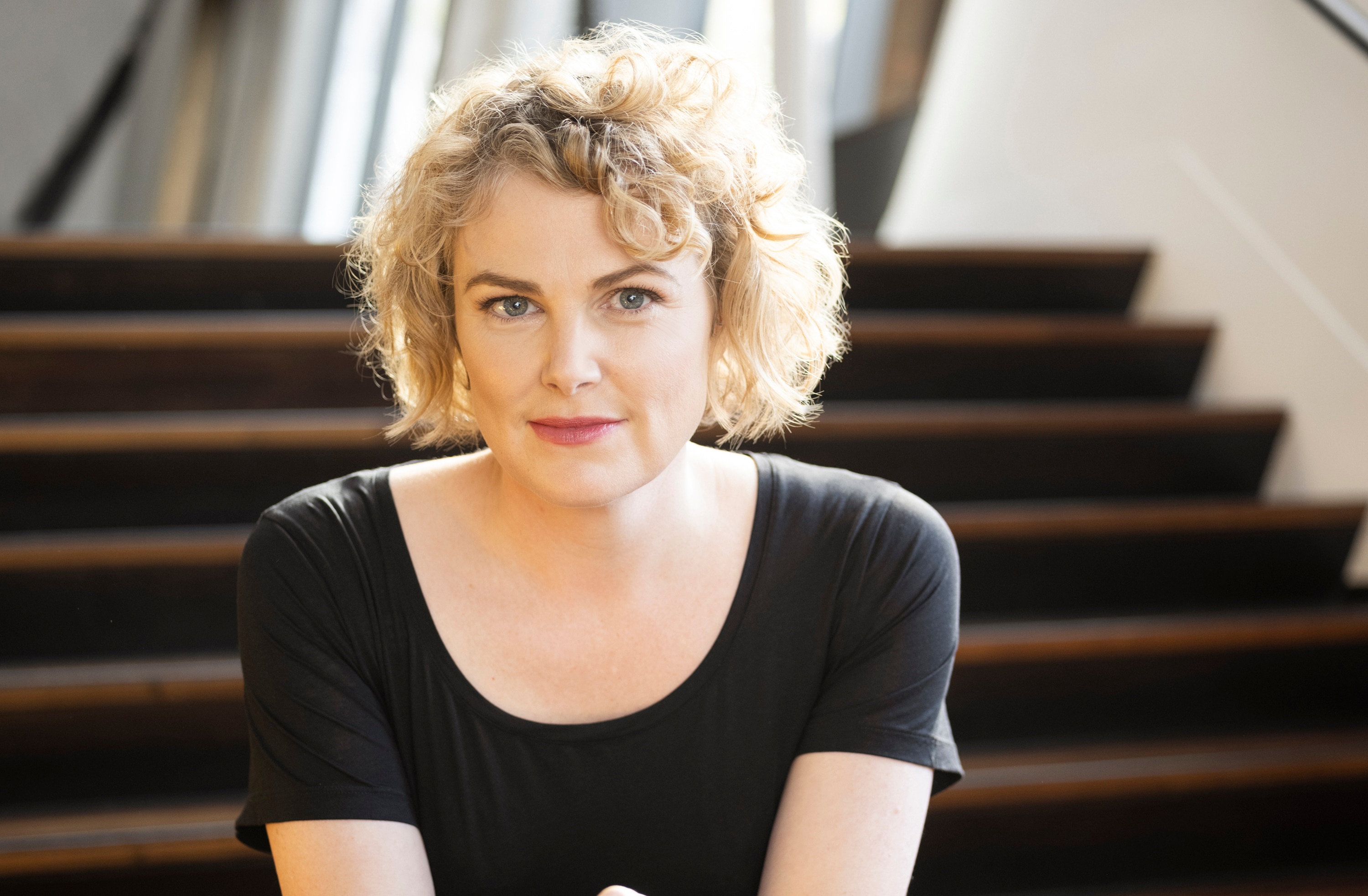 A blonde, curly-haired woman, wearing a black shirt, sits on theatre foyer stairs, with a slight smile on her face