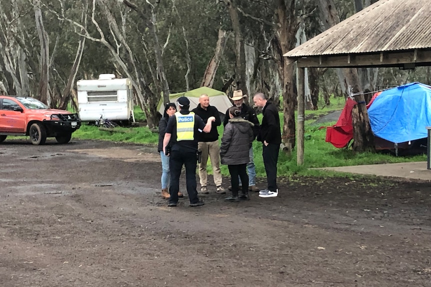 Police talk to a group of people standing in front of a caravan.