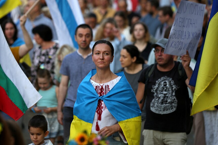Темноволосая женщина стоит посреди толпы протестующих с украинским флагом на плечах.