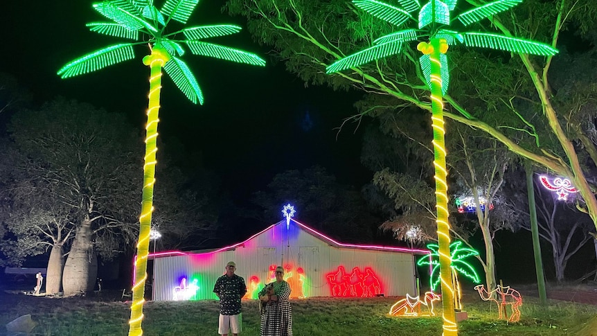 A man and a woman holding a dog stand between two palm trees lit up with lights in front of a large white shed.