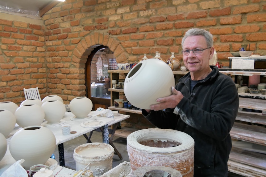 An older man with glasses stands inside a studio holding a vase in front of a pottery wheel