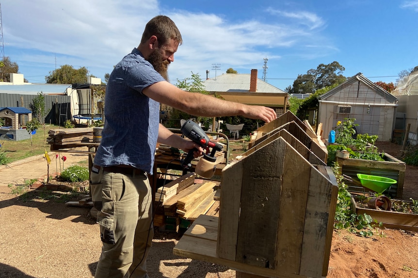 Man with beard wearing blue shirt holds a spray gun in front of a row of birdhouses.