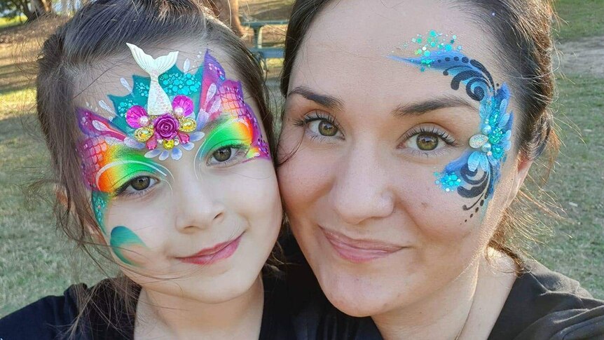 A young girl and her mother are smiling at the park, with colourful paint on their face.