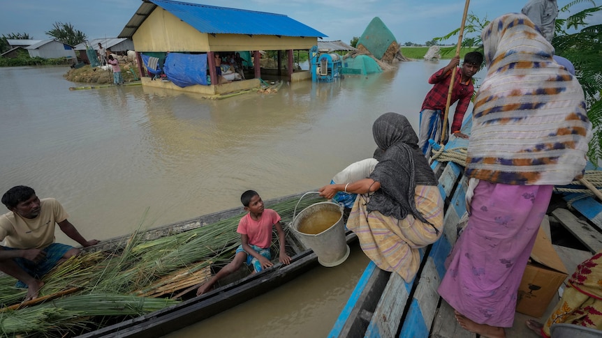 Women and children move from one canoe to another on top of a flooded farm area with a house visible