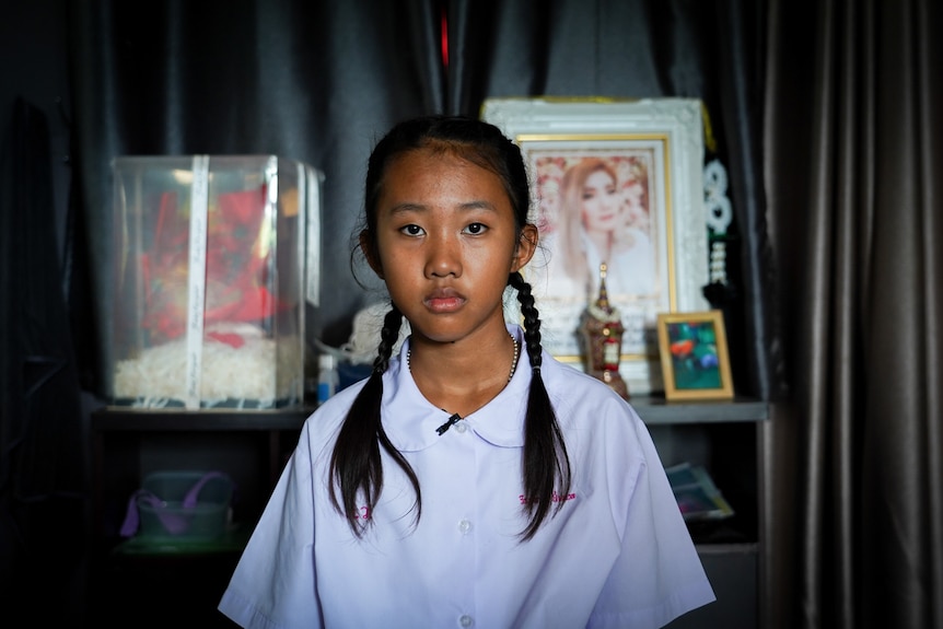 A close up of a girl with plaits sitting on a chair in front of a portrait of her mother.