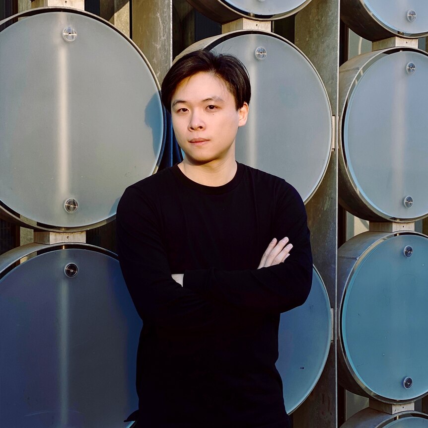 Man with short black hair, in black shirt with arms folded in front of large round metal objects. 