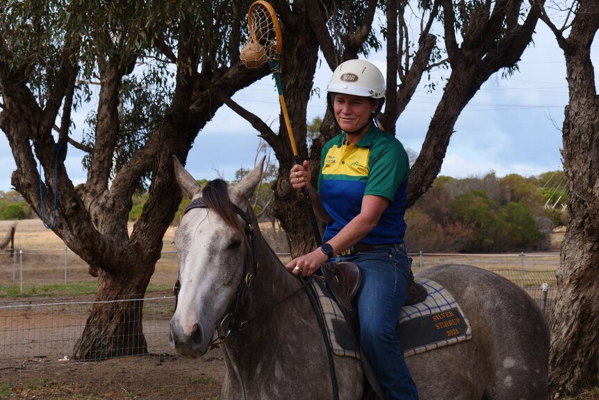 A woman rides a dappled grey horse one-handed, holding a polocrosse stick, outside. She wears blue jeans and a helmet.