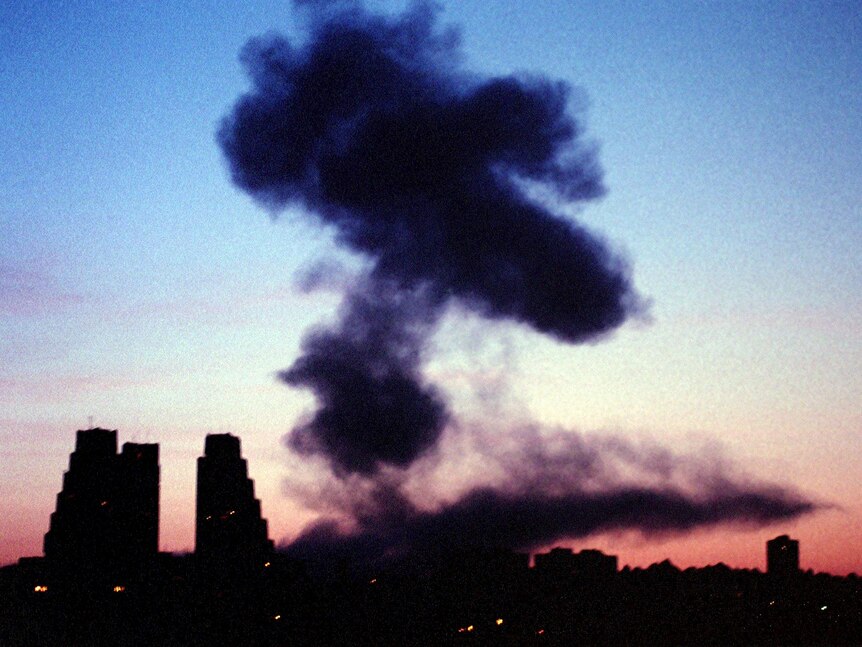 A puff of black smoke rises in the air in front of some buildings in the twilight