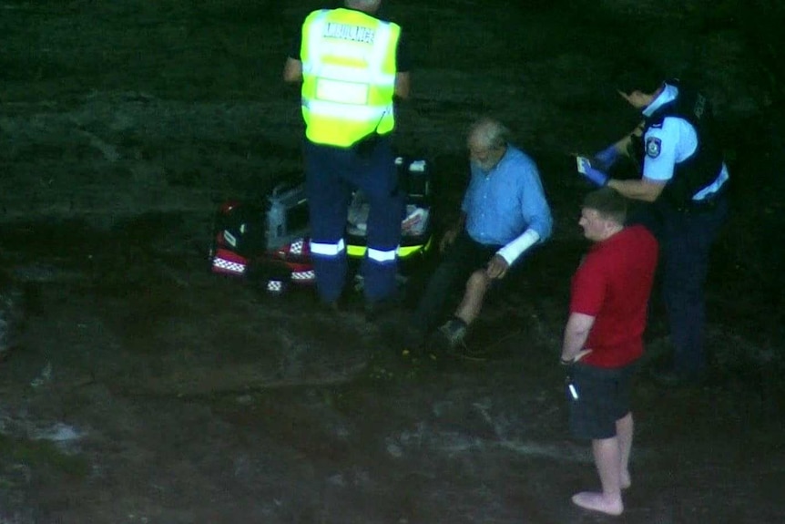 A man sits on a rock face as paramedic wraps a bandage around his wrist.