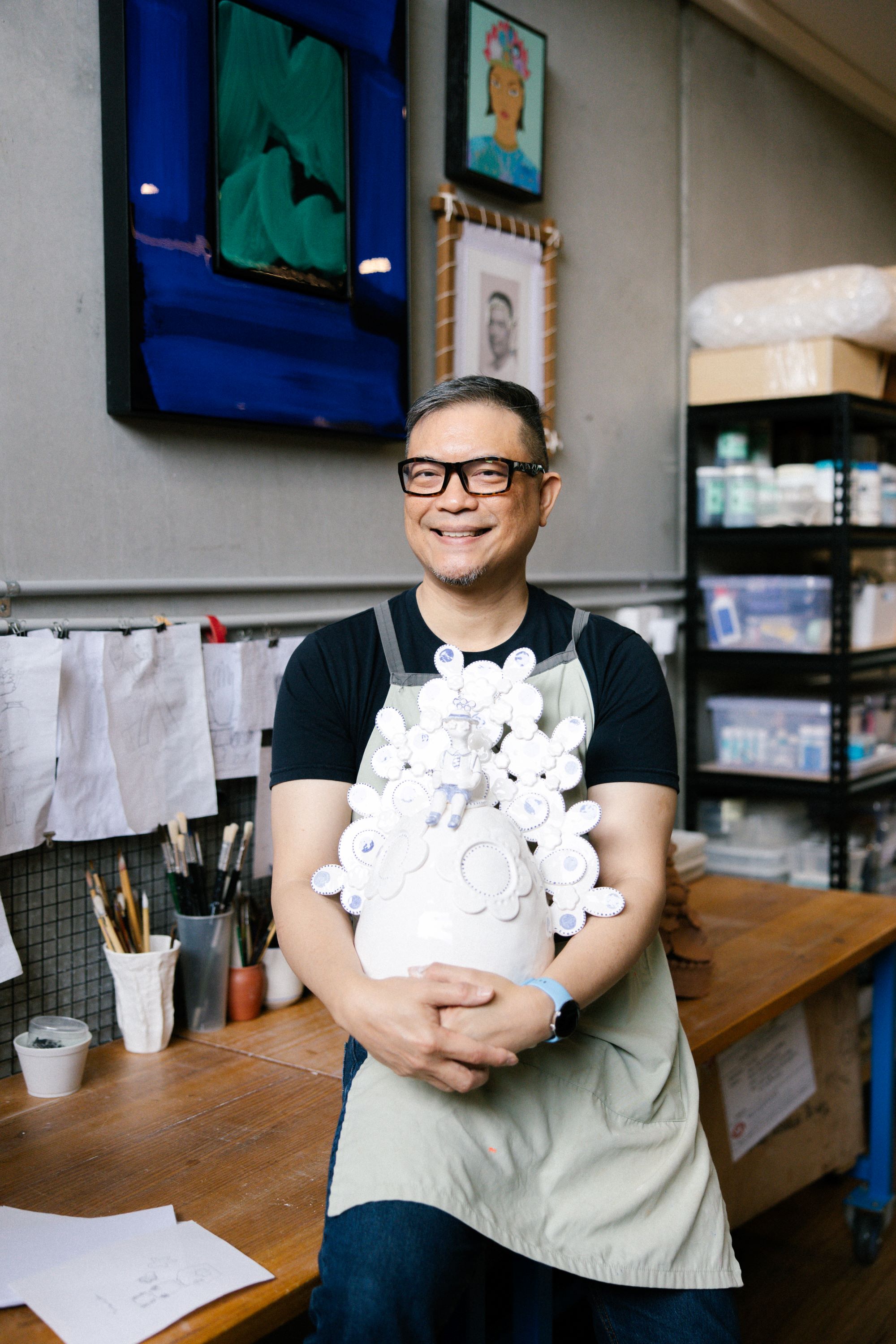An Asian man with short greying hair and glasses, holding a white ceramic sculpture, sits on the edge of a table