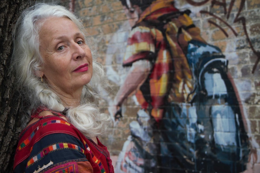 Sarah Jane Adams stands leans against a tree truck in front of a brick wall mural.