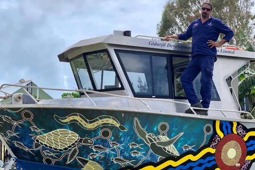 A man stands on a boat that has Aboriginal Artwork on the hull