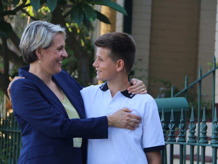 A woman with short blonde hair hugging her teenage son in front of the front gate to a house.