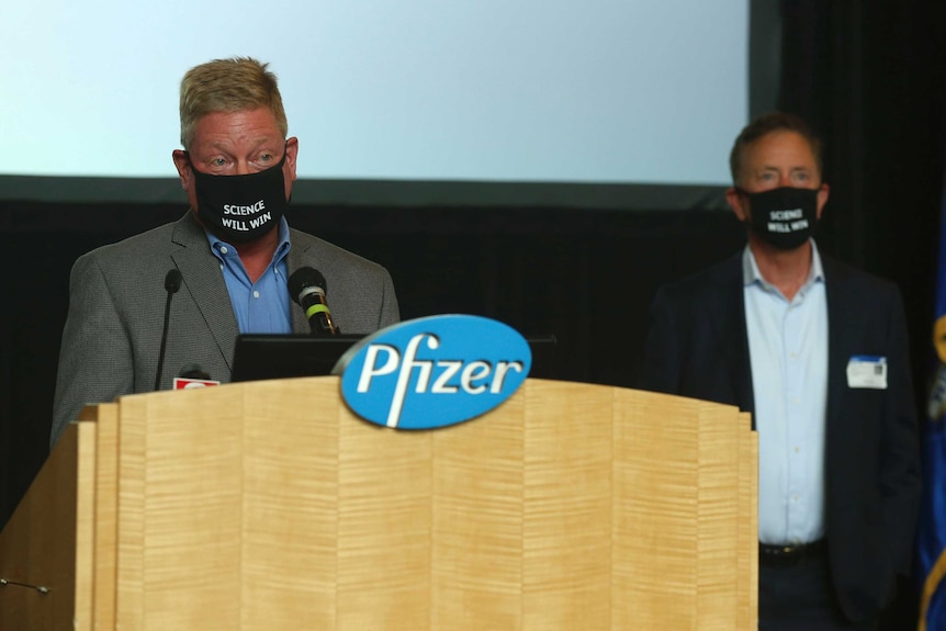 Two men speak from a podium wearing face masks that say 'Science will win'.