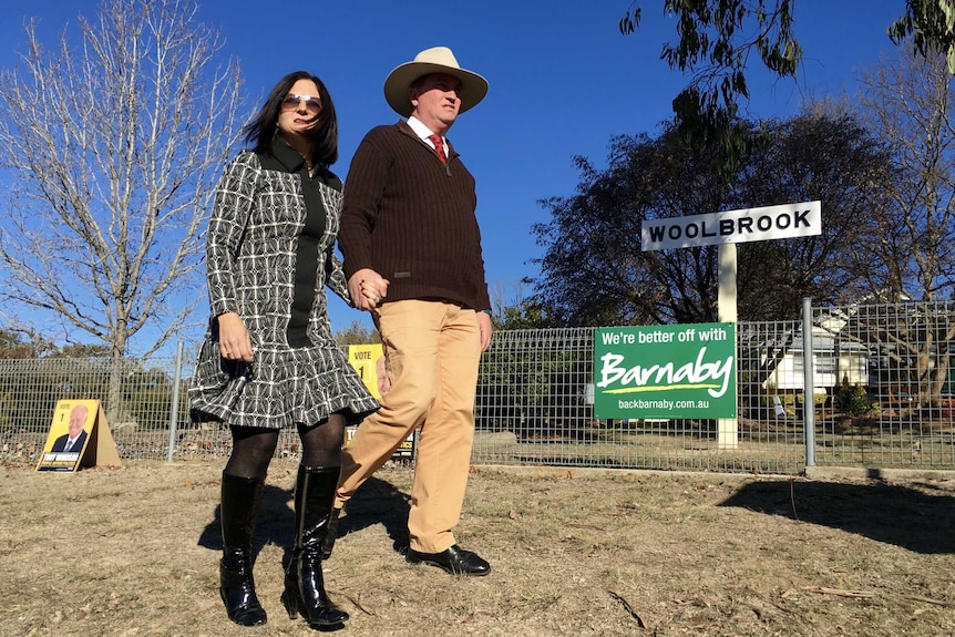 Barnaby and Natalie Joyce hold hands while walking past a 'Woolbrook' sign.