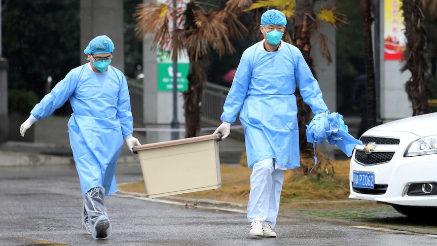 two people in blue medical gowns hair netting and face masks carry a tub along a road outside near a car