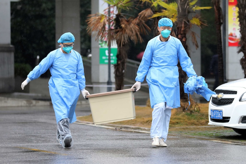 Two people in blue medical gowns hair netting and face masks carry a tub along a road outside near a car