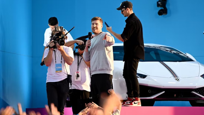 A man stands on stage pointing to a crowd while holding a microphone, a luxury car behind him