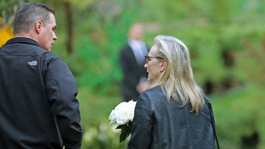 Meryl Streep holds white flowers and talks a man while walking.