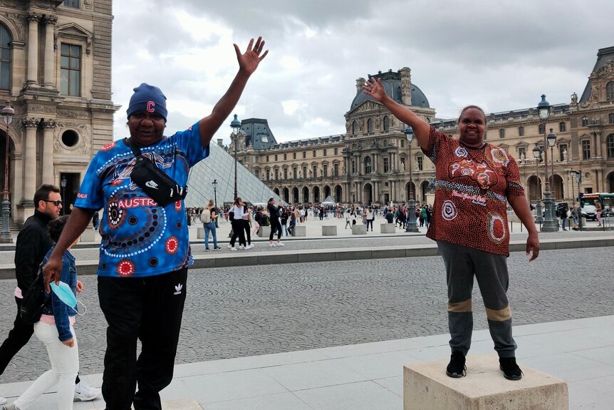 two Indigenous woman wearing colourful t-shirts standing on blocks with their arms raised in front of the Louvre in Paris