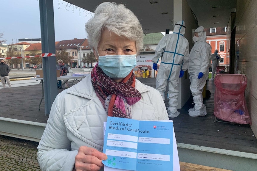 An older woman with short grey hair in a face mask holding a medical certificate