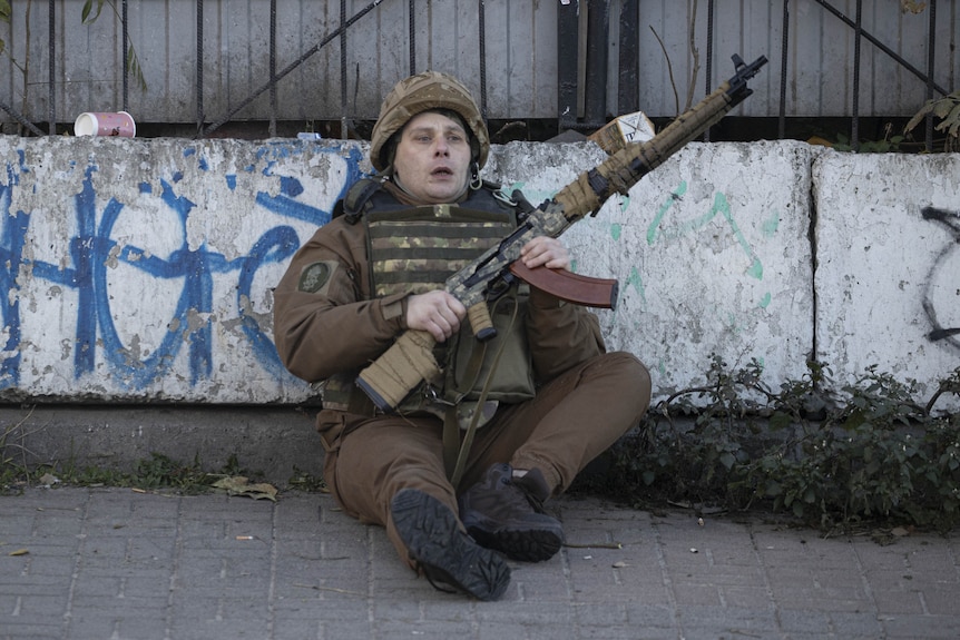 A soldier sits on the ground holding a gun and wearing military protective wear. His face is pale and mouth open in shock. 