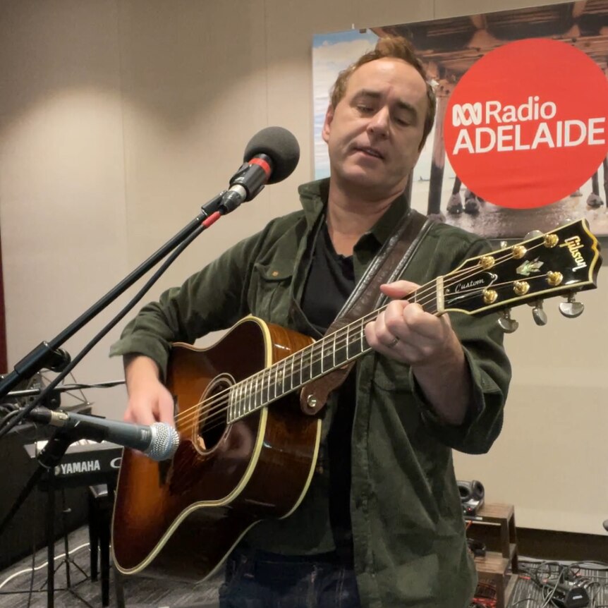 man playing guitar behind microphone in radio studio with signage in background