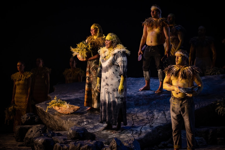 Five Indigenous performers wearing yellow ochre and some holding wattle, on stage, singing.