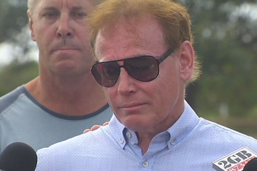 An emotional man with fair hair, wearing dark sunglasses, stands outside and speaks to the media.