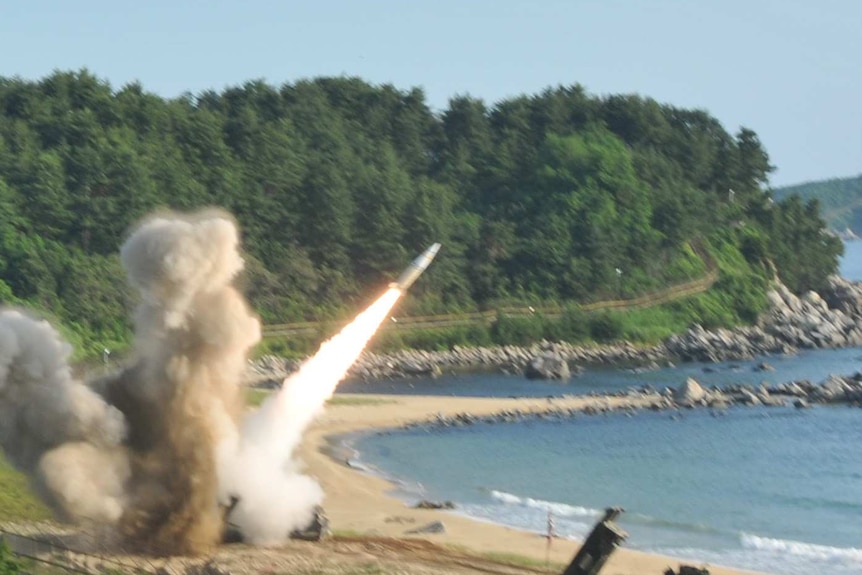 A missile is fired on a beach. Lush green forestry is the the background of the rocket blasting into the air over the ocean
