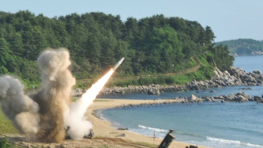 A missile is fired on a beach. Lush green forestry is the the background of the rocket blasting into the air over the ocean