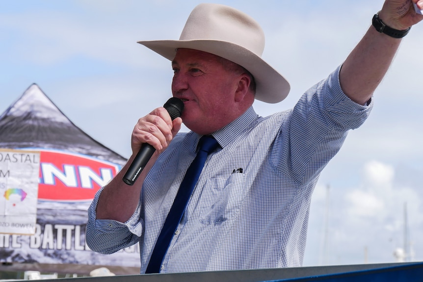 A man wearing a cowboy hat speaking into a microphone with his hand up in the air. 