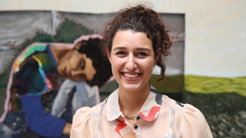 A young woman with curly dark hair pulled into a ponytail smiles in front of a large textile artwork.