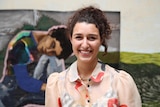 A young woman with curly dark hair pulled into a ponytail smiles in front of a large textile artwork.