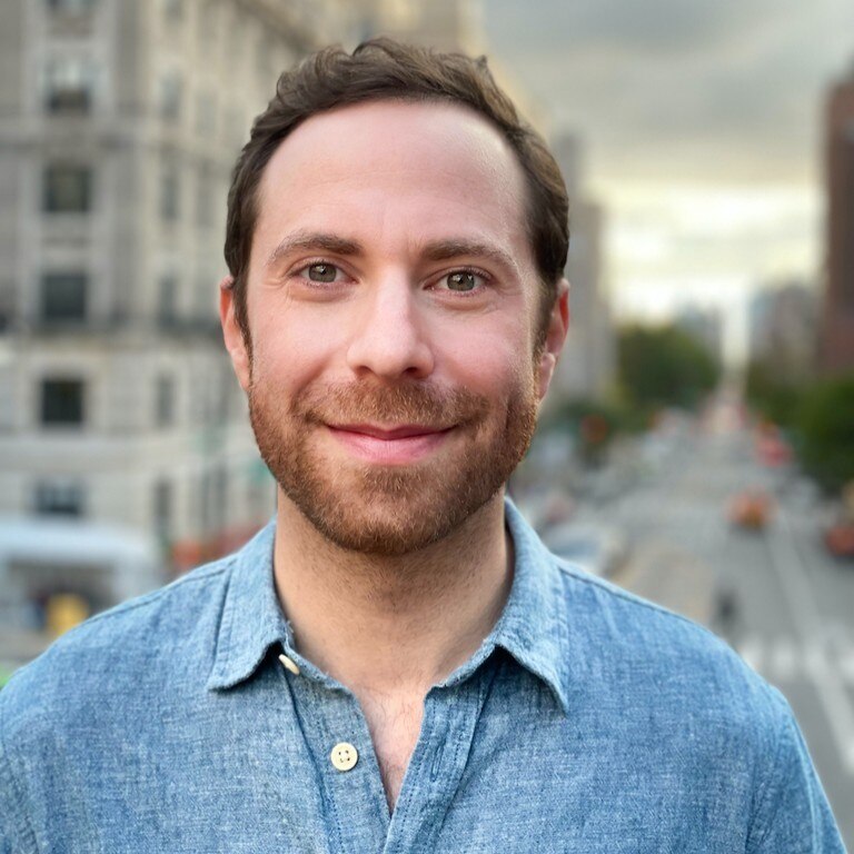 Un hombre con cabello castaño corto y una camisa azul con botones sonríe.