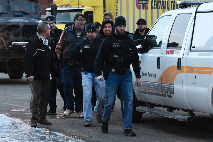 A group of at least 10 men wearing winter clothing walk along the road escorted by a police officer, with vehicles nearby.