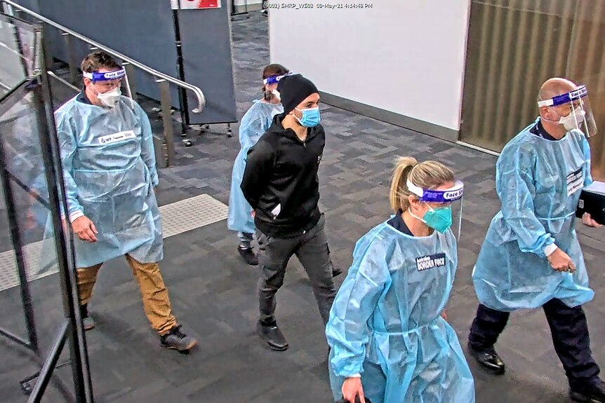 A young man in dark clothing and a surgical face mask, is escorted through an airport by four people in protective equipment.